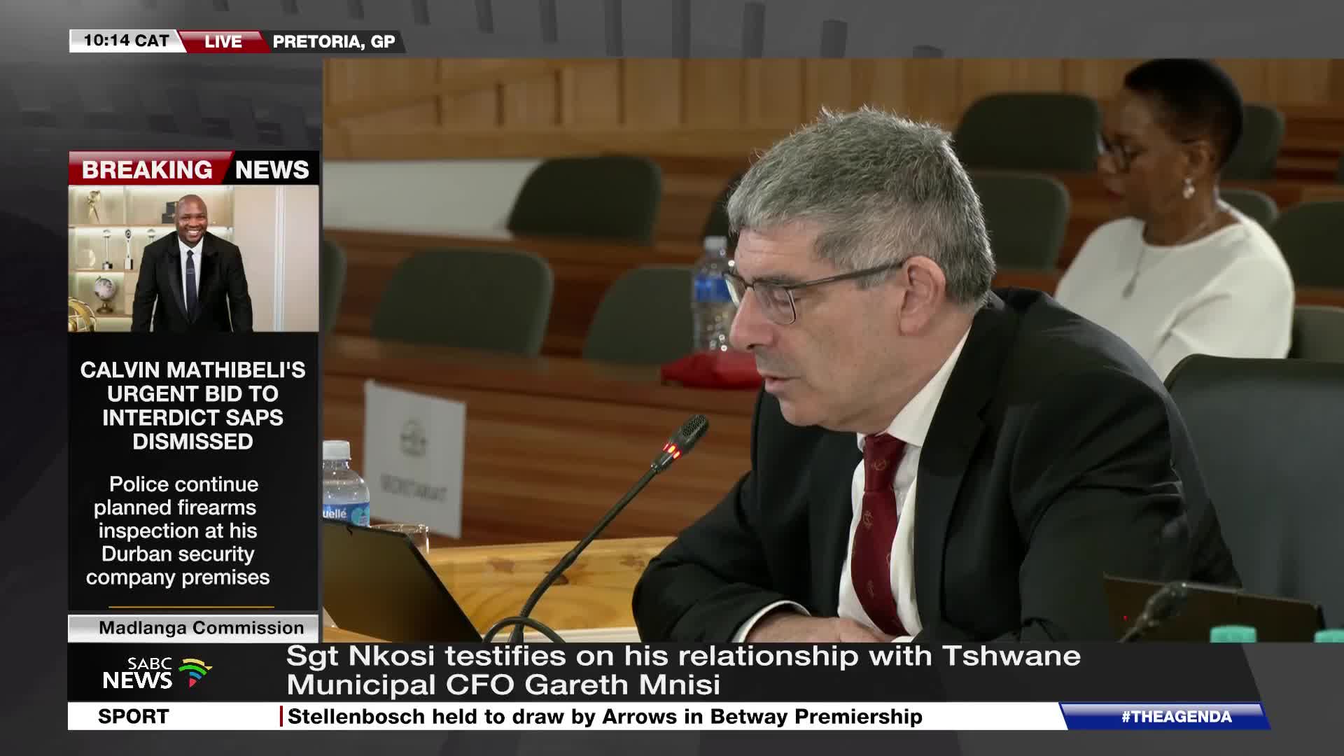 A man in a suit and tie leans into a microphone, speaking at a commission hearing. The SABC News chyron below identifies him as Sgt Nkosi, testifying about his relationship with a municipal CFO.