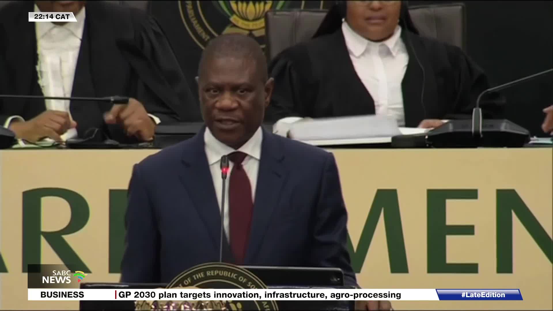 A man in a dark suit and red tie speaks at a podium, his voice amplified by a microphone. Behind him, individuals in formal robes sit, their hands resting on the table. The SABC News chyron at the bottom indicates this is a broadcast from South Africa.