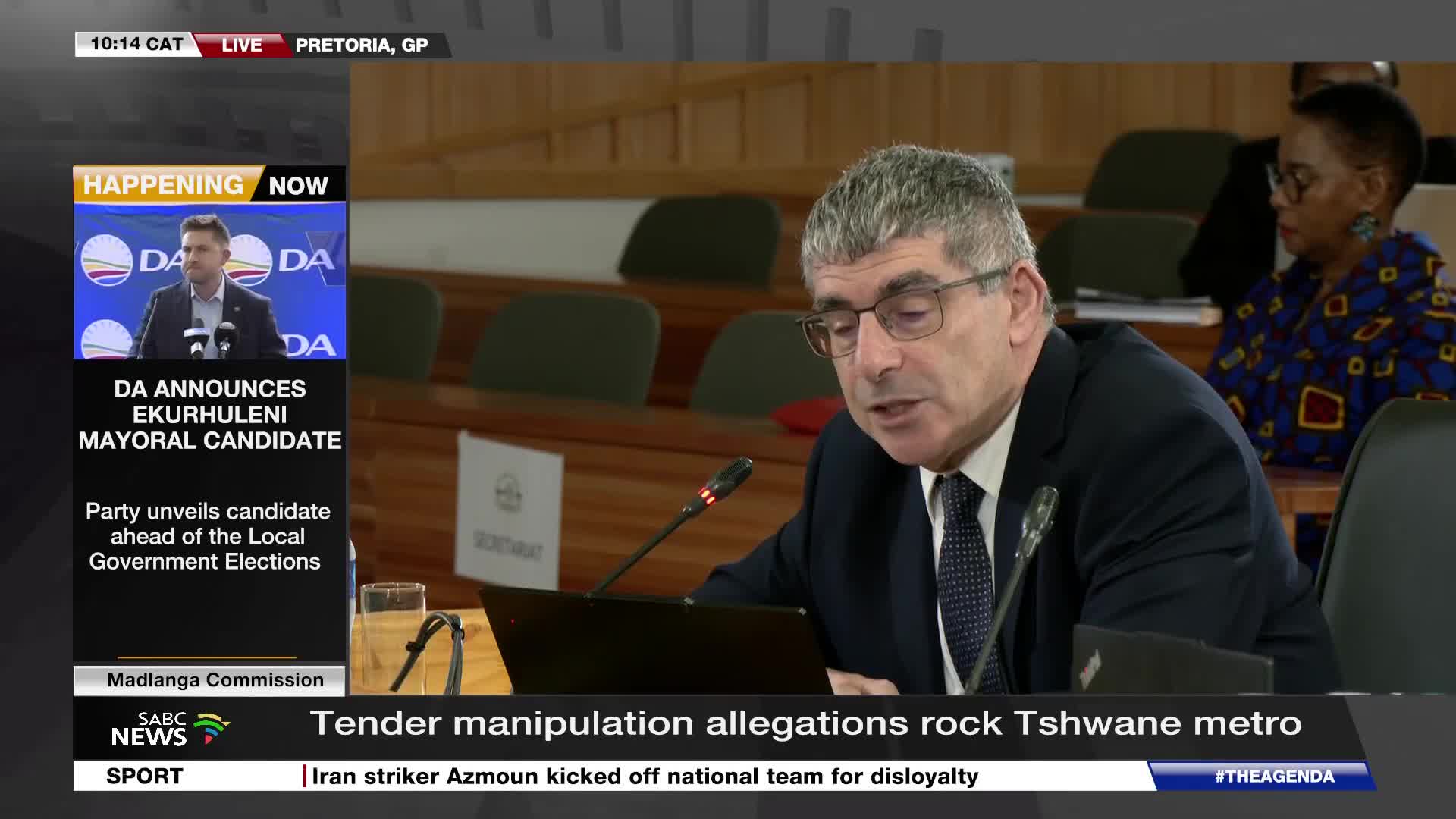 A man in a suit speaks into a microphone at a table, while SABC News reports on tender manipulation allegations. Another screen shows a politician addressing a crowd, announcing the DA's Ekurhuleni mayoral candidate.