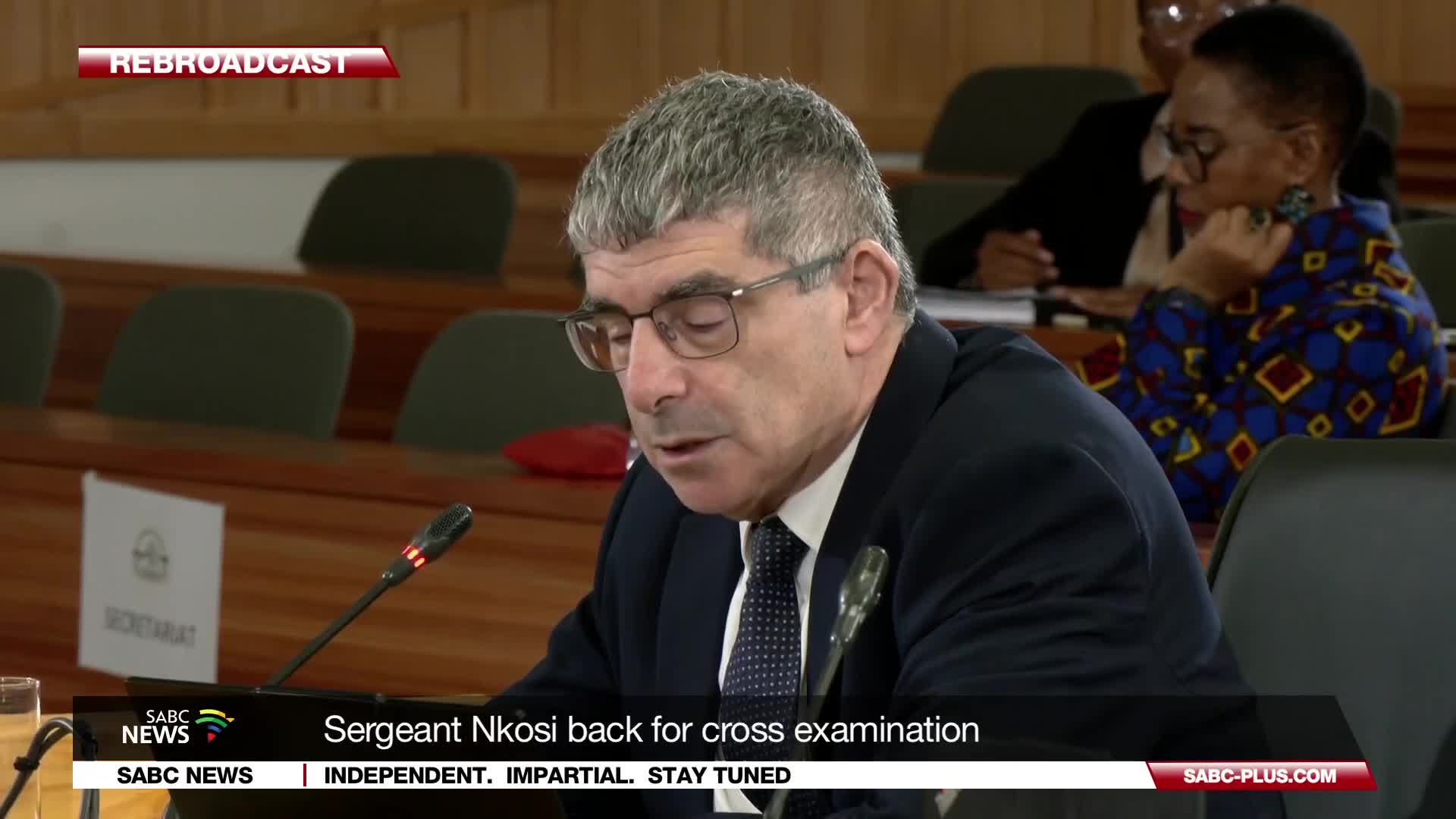 A man in a suit and tie speaks into a microphone, his glasses reflecting the light. Behind him, a woman in a vibrant blue and yellow patterned jacket listens intently. This is a SABC News broadcast from South Africa, showing Sergeant Nkosi back for cross-examination.