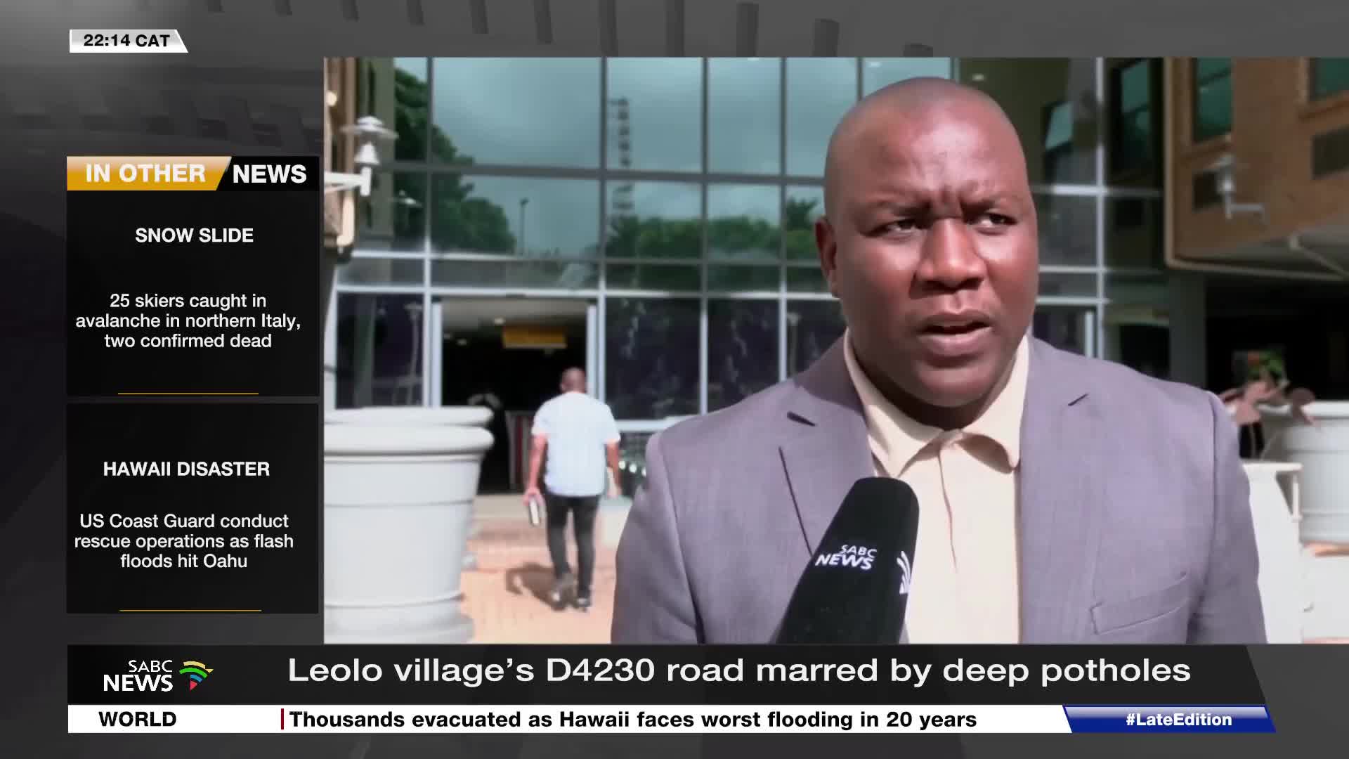 A man in a suit speaks into an SABC News microphone, while behind him, another man walks away from the camera into a building. The screen displays news headlines from South Africa and around the world.