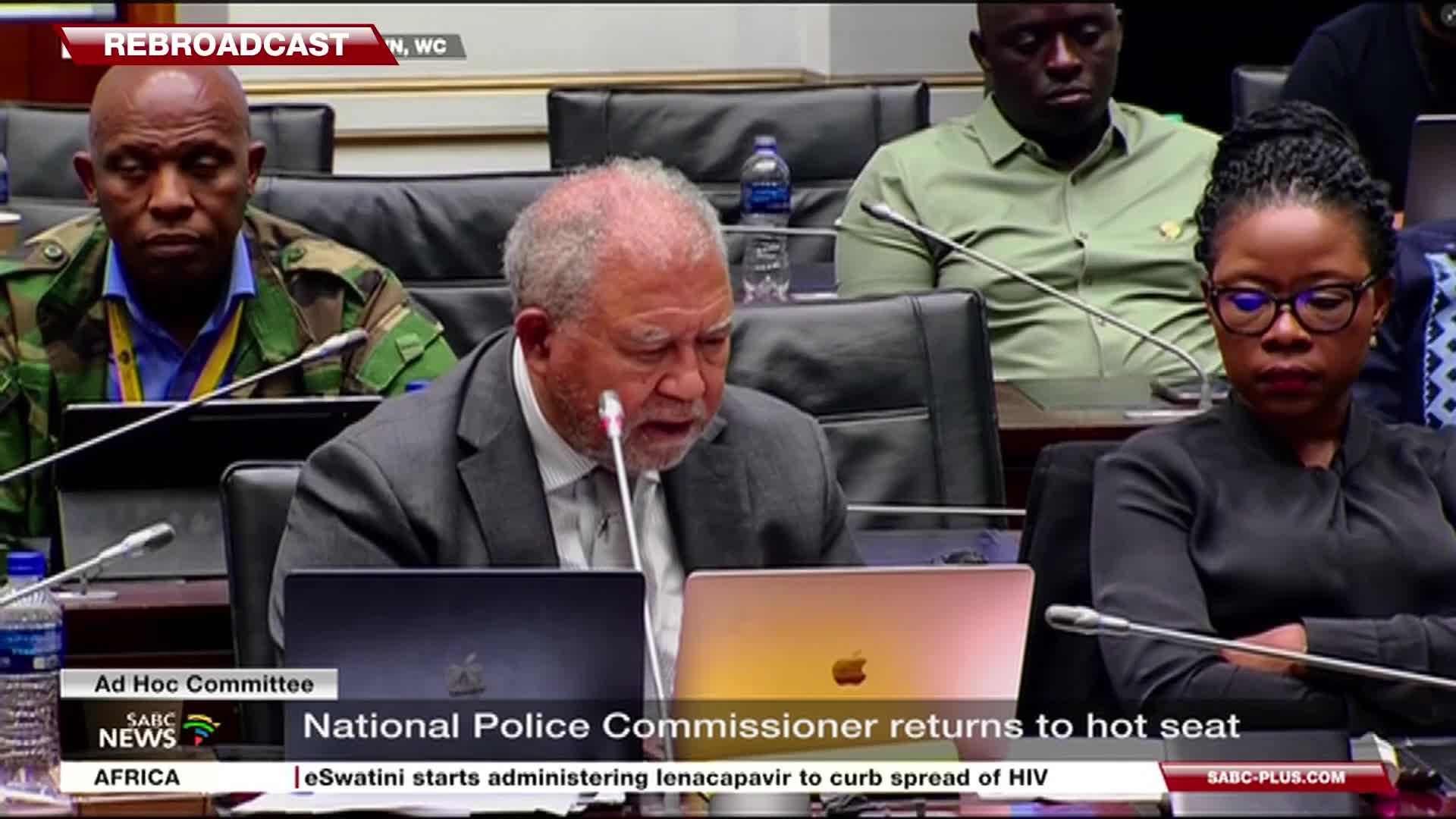 The National Police Commissioner, a man with a grey beard and suit, leans into a microphone at a table. He's speaking at a hearing, with other officials seated around him, their faces serious.