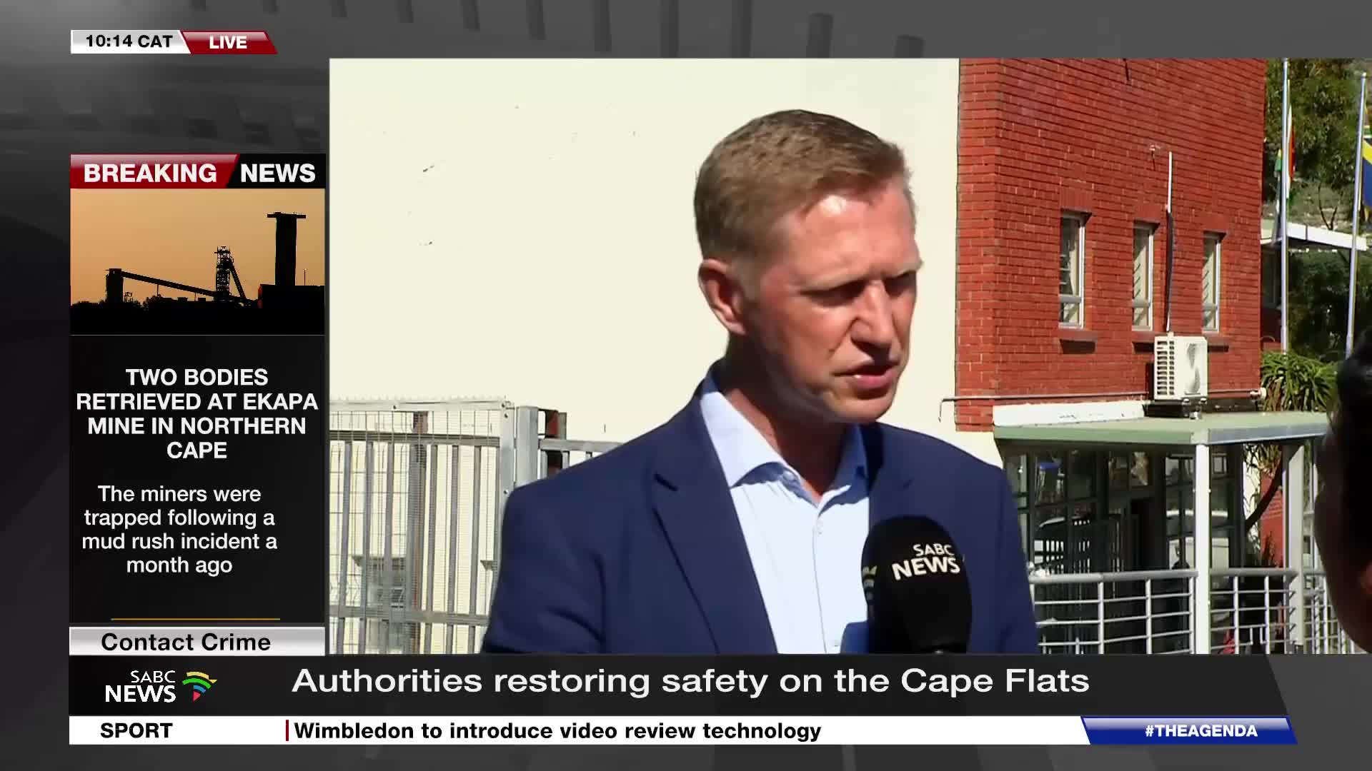 A man in a blue suit speaks into an SABC News microphone. Behind him, a brick building stands under a bright sky.