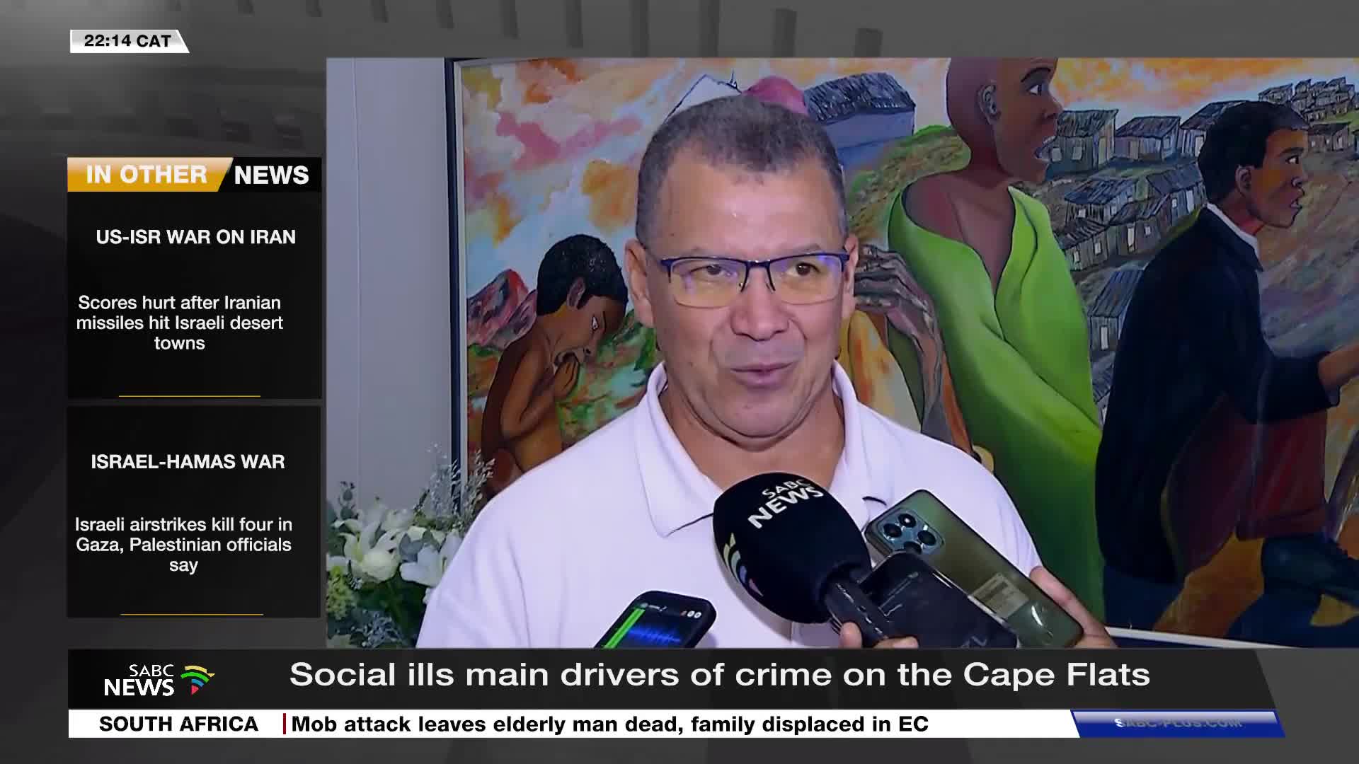 A man in a light pink shirt is speaking into an SABC News microphone. Behind him, a vibrant mural depicts figures against a backdrop of informal housing.