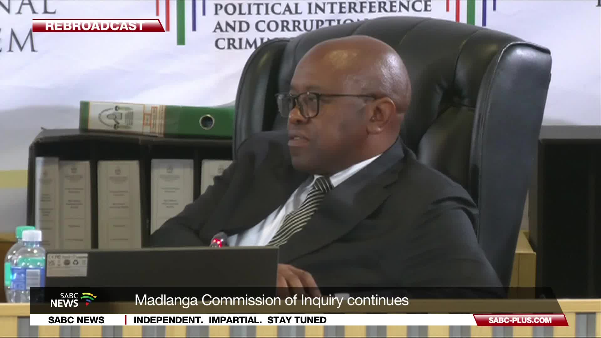 A man in a suit and tie sits in a large chair, listening intently. Behind him, shelves are stacked with thick, bound documents, suggesting the ongoing proceedings of the Madlanga Commission of Inquiry.