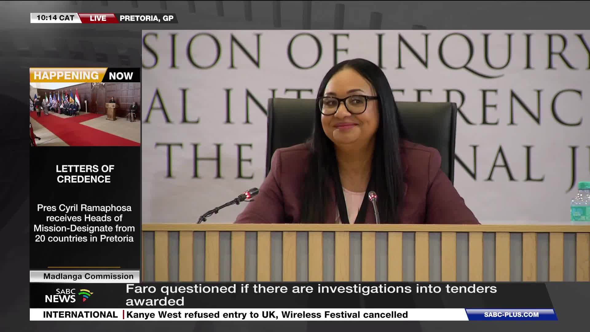 A woman with glasses and dark hair smiles faintly as she sits behind a wooden desk. On the screen, SABC News reports on a commission of inquiry and President Ramaphosa receiving diplomats.