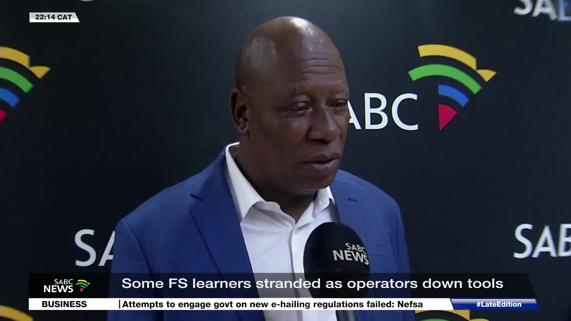 A man in a blue suit speaks into an SABC News microphone. The background displays the SABC logo and text about stranded learners.