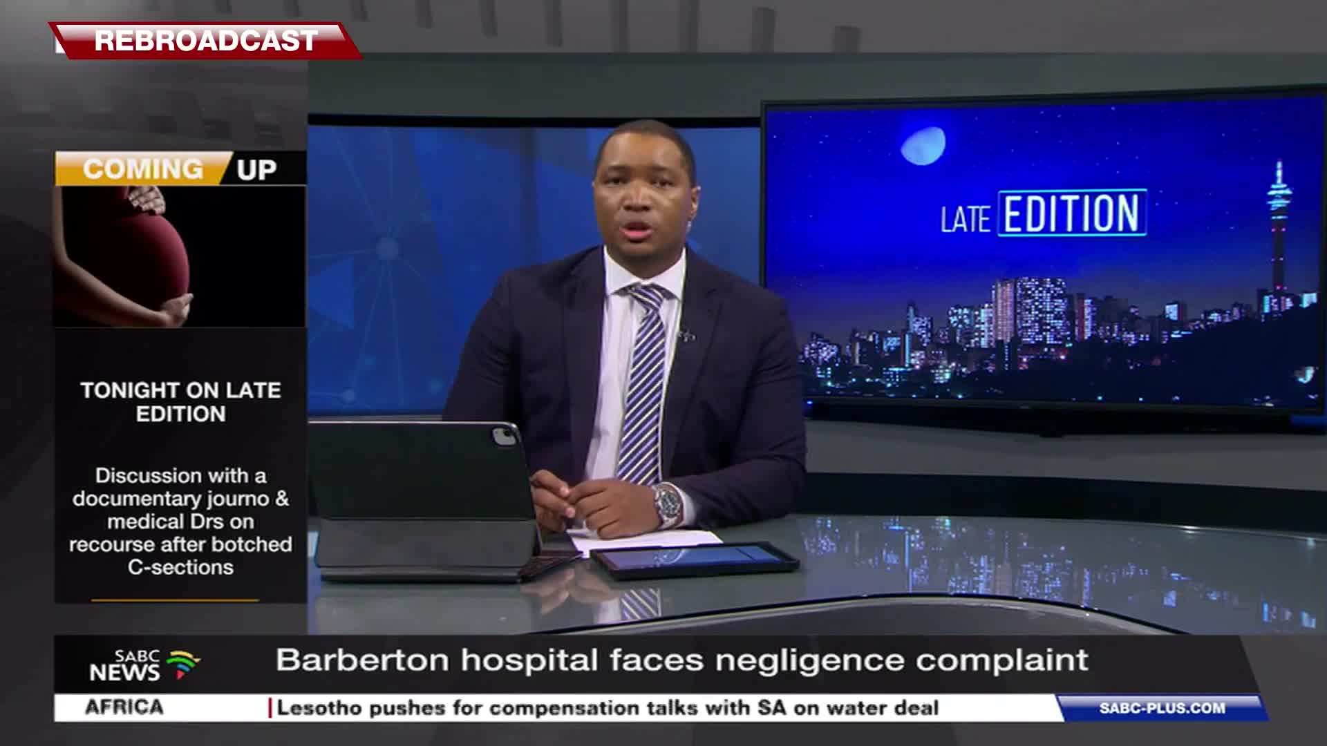 A news anchor sits at a desk, speaking into a microphone. Behind him, a large screen displays the SABC News logo and a city skyline at night.