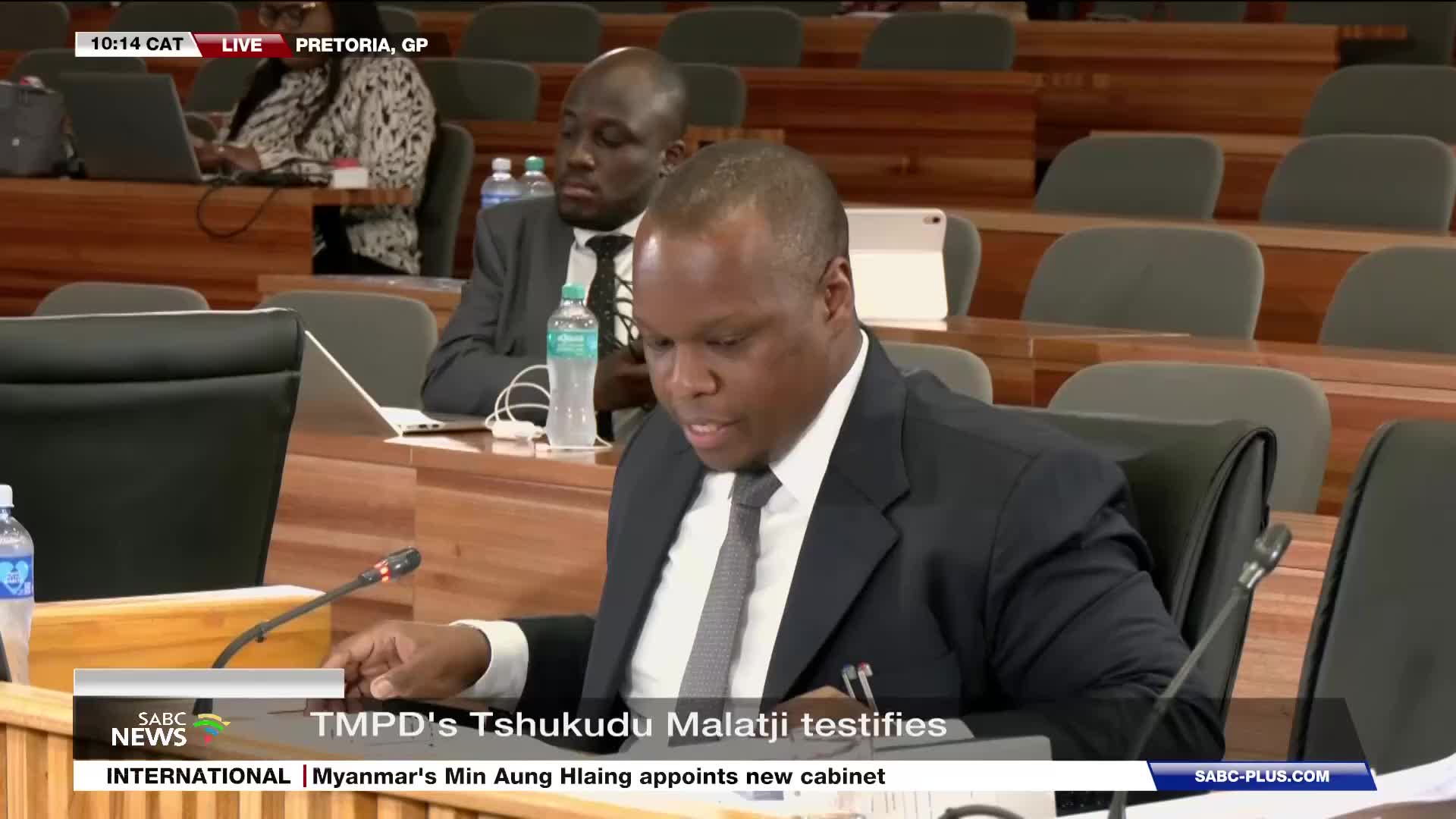 A man in a dark suit and tie leans forward, speaking into a microphone. Behind him, other individuals sit at tables in a room with rows of empty chairs.