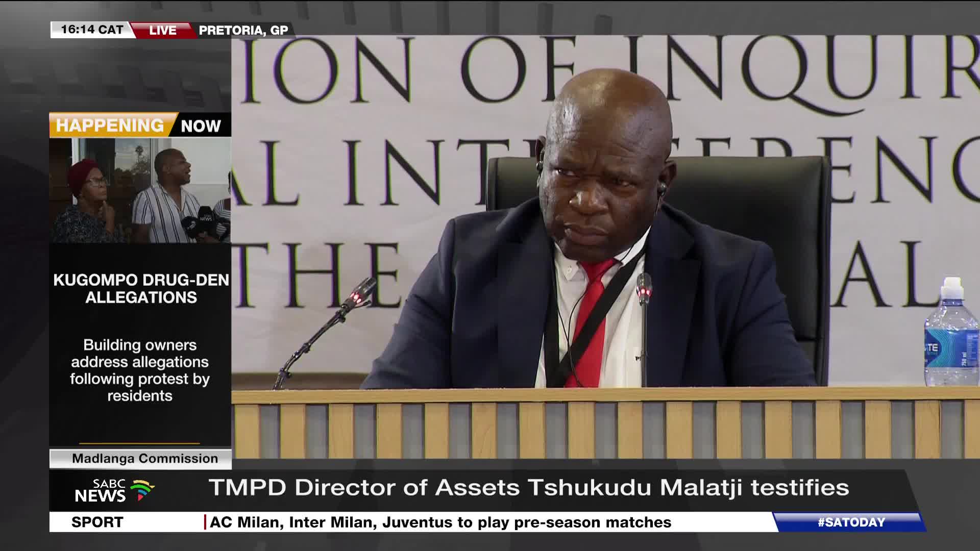 A man in a dark suit and red tie sits at a table, speaking into a microphone. Behind him, a banner reads "COMMISSION OF INQUIRY".