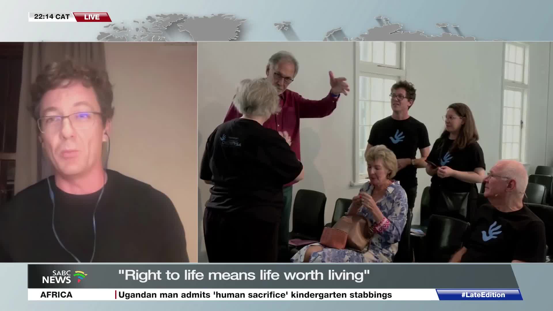 A man in a maroon shirt gestures animatedly while speaking to a woman in a black t-shirt. Behind them, others in similar black shirts stand and sit, some looking towards the speaker. The SABC News broadcast shows this scene live from South Africa.