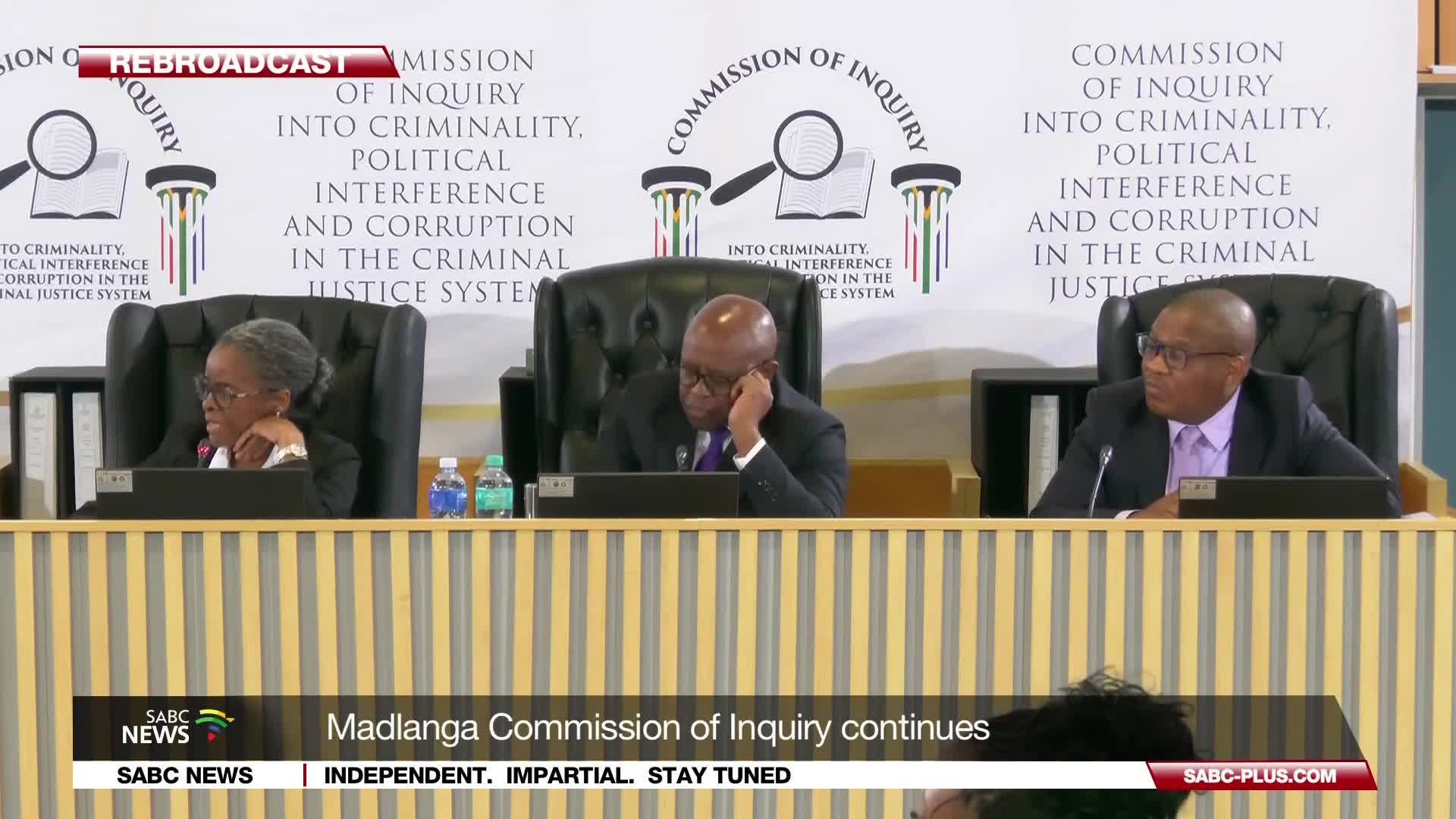 The Madlanga Commission of Inquiry continues its proceedings as three commissioners sit behind a wooden desk. The woman on the left rests her chin on her hand, while the man in the center leans forward, head bowed, and the man on the right looks towards the side.
