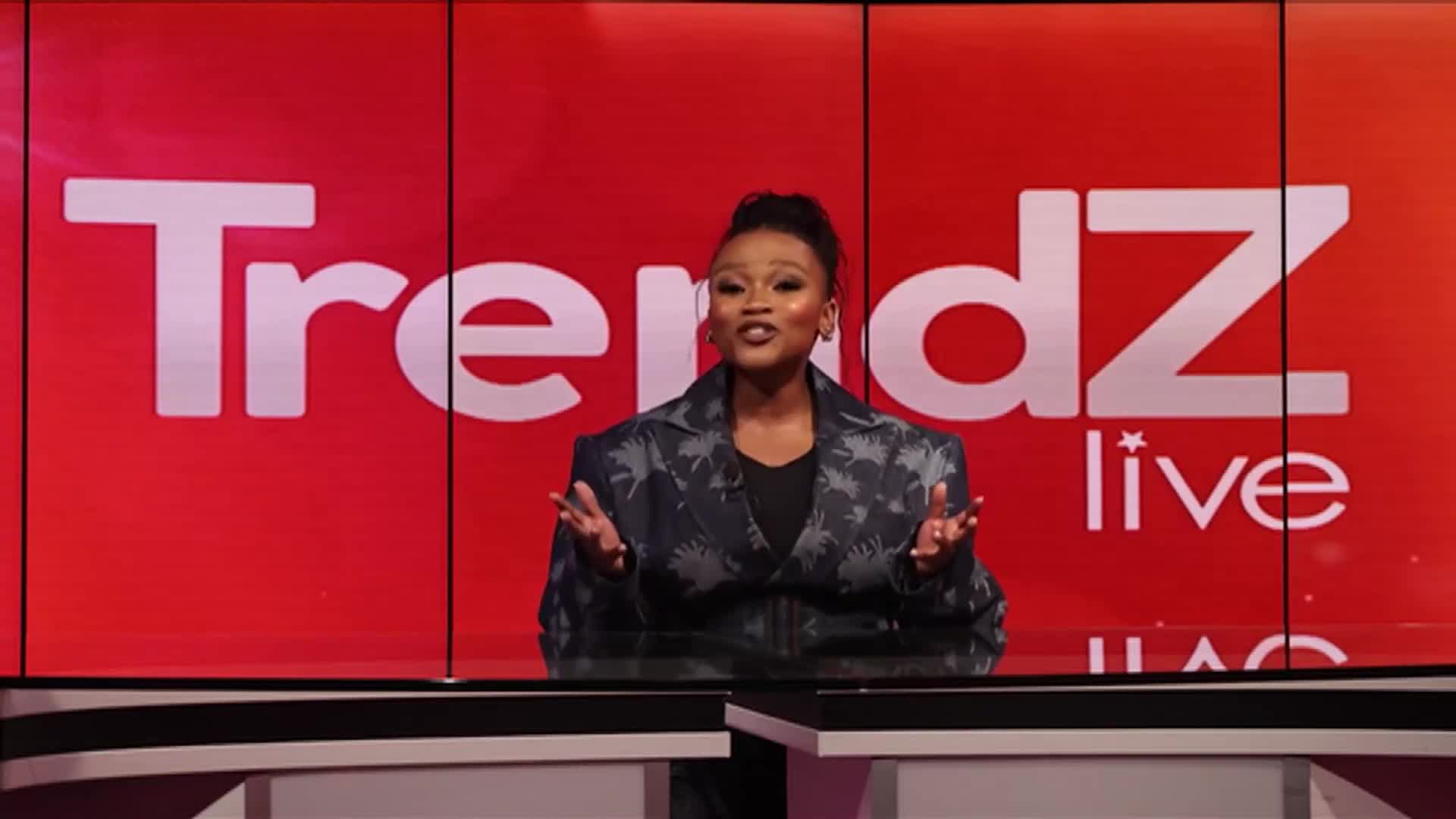 A woman stands behind a desk, her hands outstretched as she speaks. Behind her, the SABC News logo "Trendz live" glows in large white letters against a vibrant red screen.