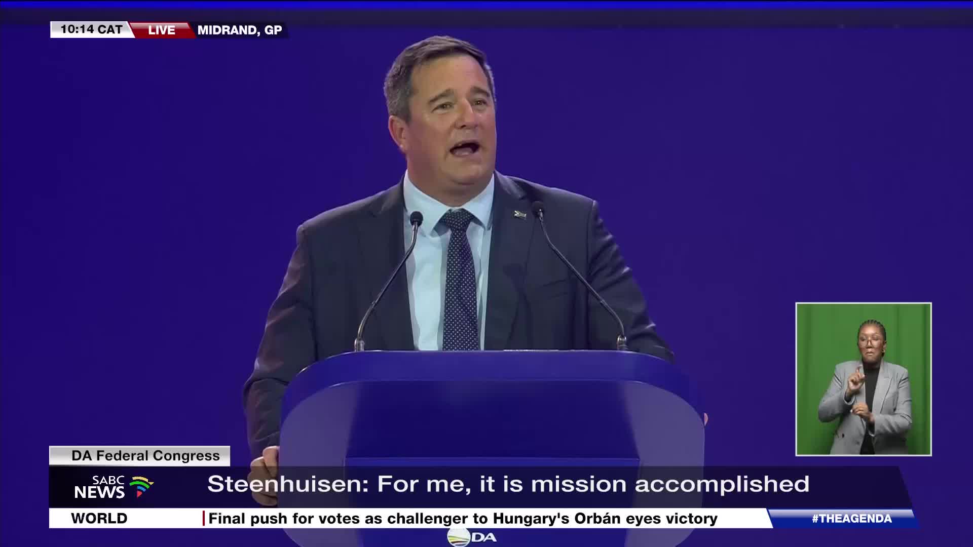 A man in a dark suit speaks from behind a podium, his mouth open. To the right, a woman in a grey jacket signs with her hands.