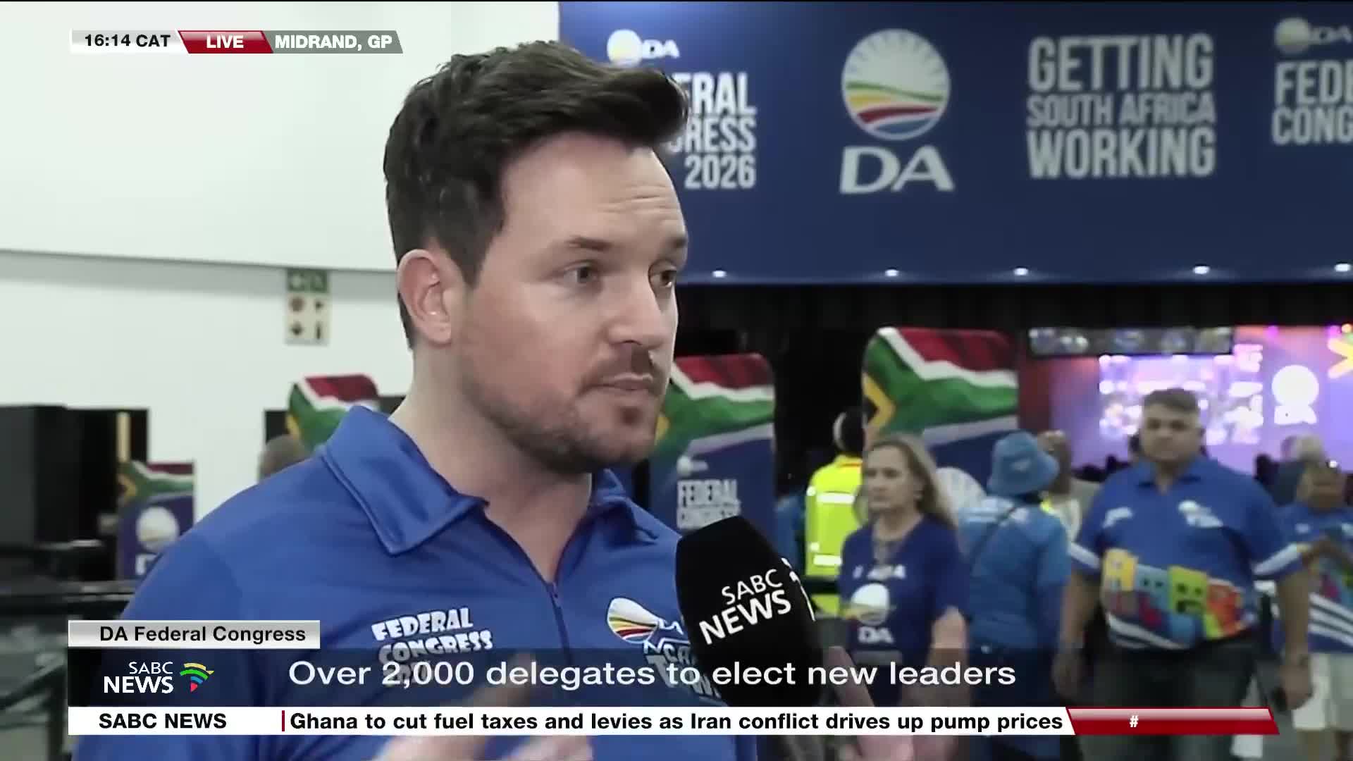 The SABC News reporter stands before a backdrop for the DA Federal Congress, with delegates milling about behind him. A large banner overhead reads "GETTING SOUTH AFRICA WORKING."