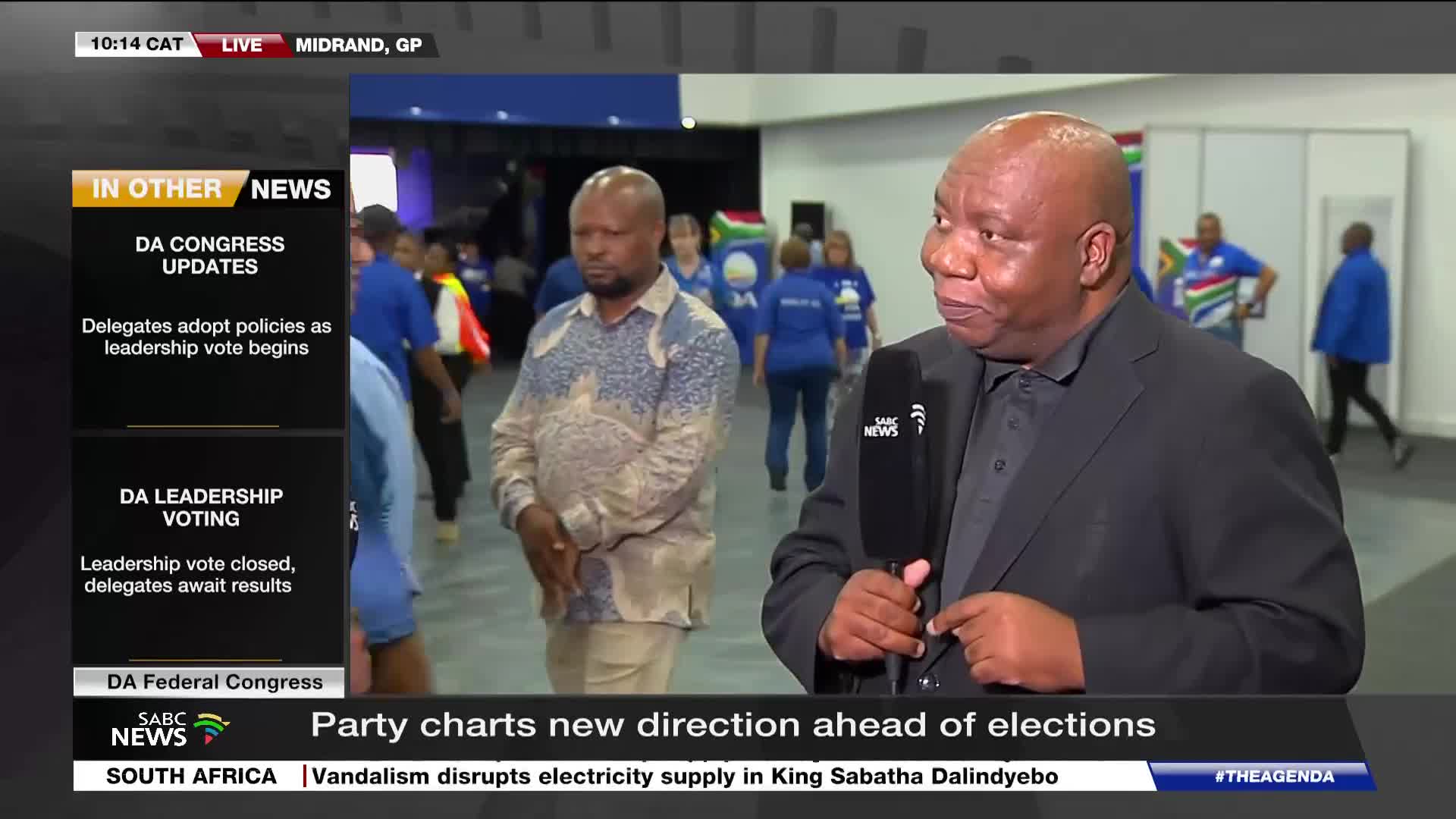 A man in a black suit holds an SABC News microphone, speaking to the camera. Behind him, people in blue shirts move through a hall, some carrying South African flags.
