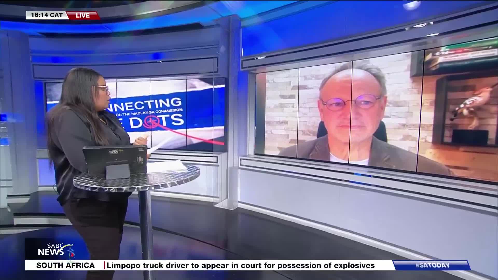 A woman in a dark shirt stands at a metal table, looking at papers while a man in glasses appears on a large screen behind her. The SABC News logo is visible, along with a headline about a Limpopo truck driver.