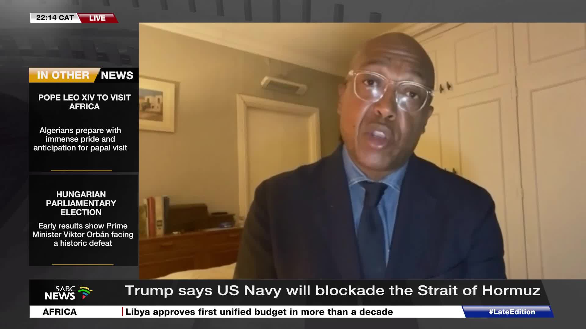 A man in a suit and tie speaks directly to the camera, his glasses reflecting the light. On the left, SABC News displays headlines about a papal visit to Africa and Hungarian election results.