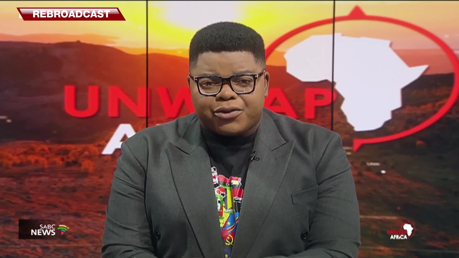 A man in a dark suit stands before a screen displaying "UNWRAP AFRICA" and a map of the continent. The SABC News logo is visible in the bottom left corner.
