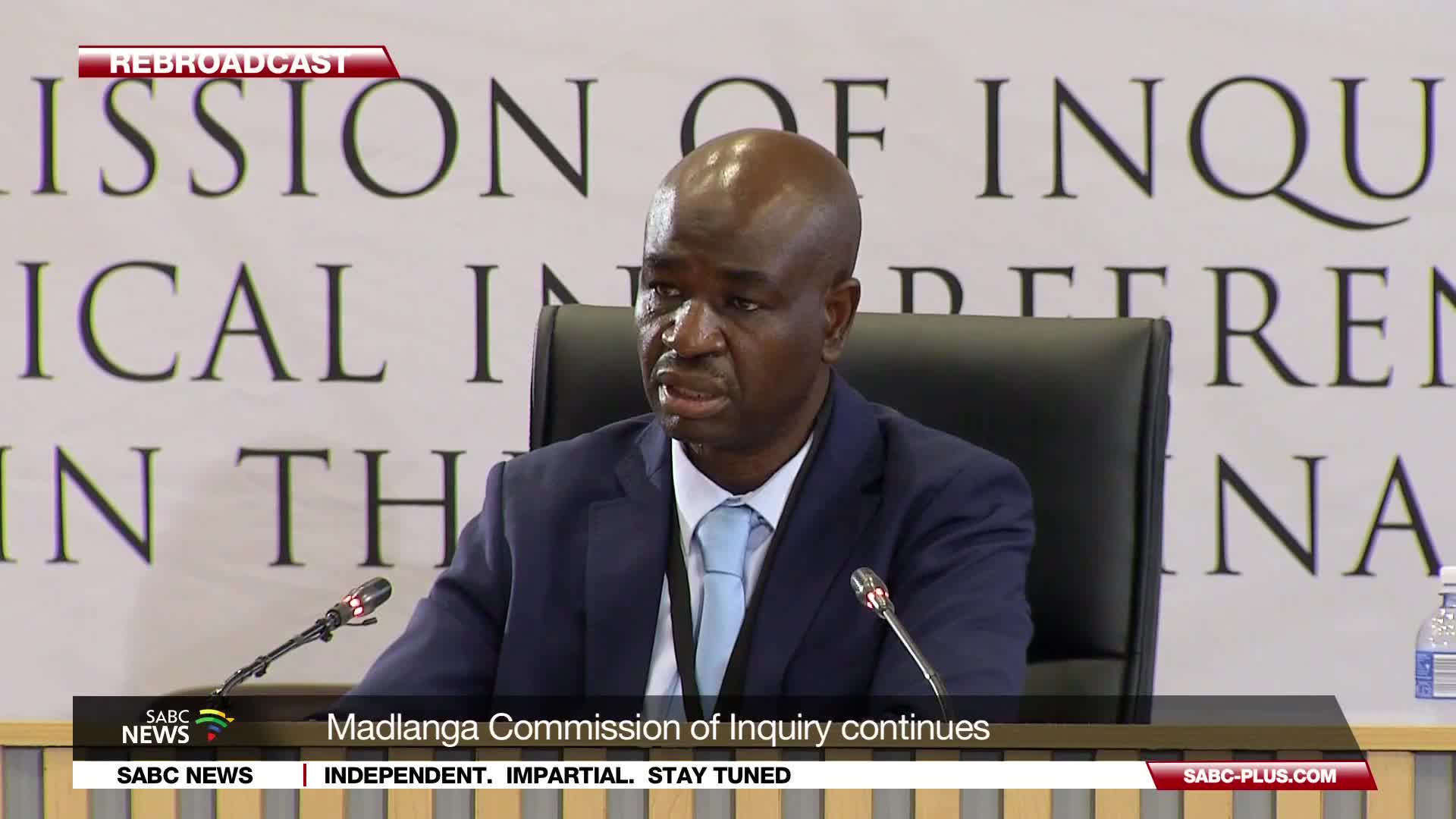 A man in a dark suit and light blue tie speaks into a microphone at a commission hearing. The SABC News banner at the bottom indicates the Madlanga Commission of Inquiry continues.
