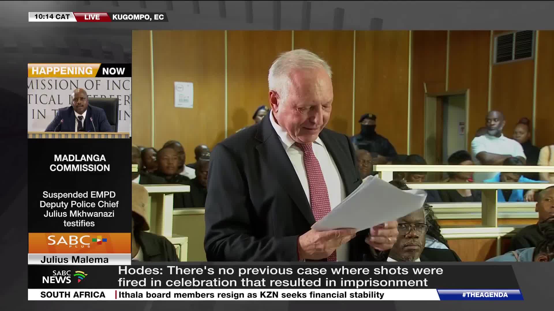 A man in a suit stands, reading from papers at the Madlanga Commission. Behind him, people are seated in what appears to be a courtroom.