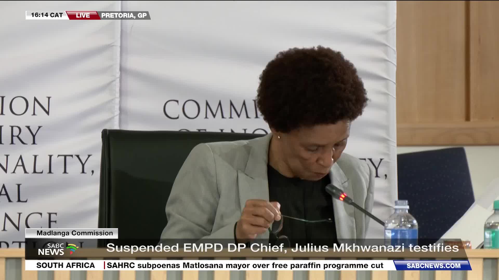 A woman in a light grey blazer leans forward, holding her glasses as she looks down. The SABC News banner at the bottom of the screen indicates this is a live broadcast from Pretoria, South Africa, where a suspended EMPD DP Chief is testifying.