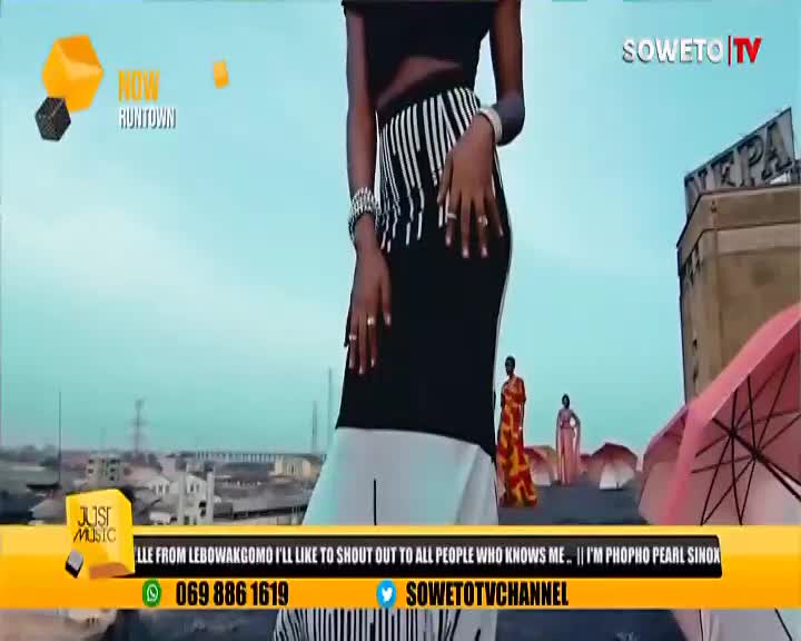 A woman in a black and white skirt stands in front of a Soweto TV backdrop, her hands gracefully posed. Behind her, two other figures stand by some open umbrellas, while a music video plays.
