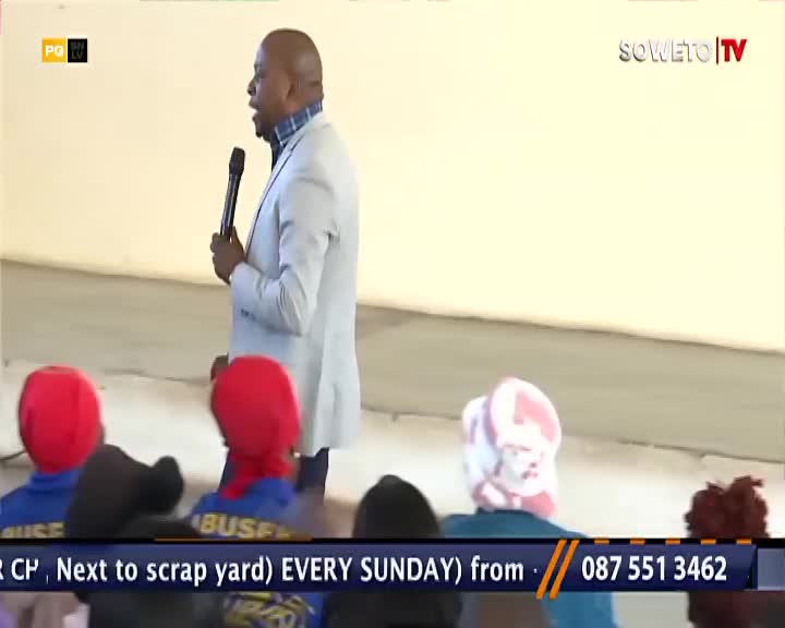 A man in a light blazer speaks into a microphone, likely on Soweto TV. In the foreground, a few people in the audience wear red and white head coverings.
