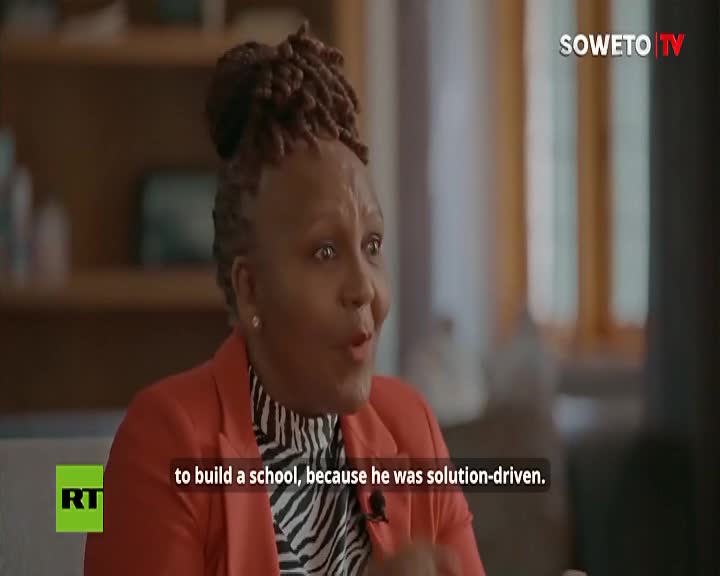 A woman on Soweto TV, wearing a red blazer over a patterned top, gestures animatedly. Her mouth is open, and the words "to build a school" are subtitled below her.
