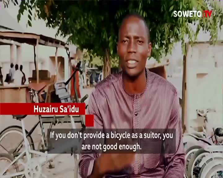 A man in a striped shirt speaks directly to the camera, with "Soweto TV" visible in the corner. Behind him, a bicycle is parked, and the caption below his name states a bold opinion about suitors.
