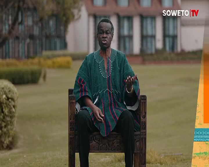 A man in a green and red patterned shirt sits on an ornate wooden chair, gesturing with his hands as he speaks. The Soweto TV logo is visible in the corner, and he's framed by a manicured lawn and a large building in the background.
