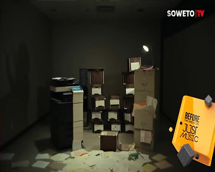 A cluttered office, maybe for Soweto TV, is dimly lit by a desk lamp. Papers are scattered across the floor, and a large copier sits next to a stack of file boxes.
