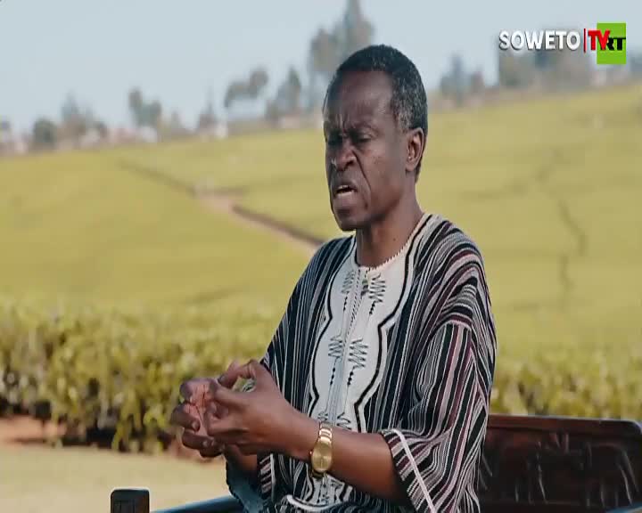 A man in a patterned shirt gestures with his hands, speaking earnestly on what looks like a Soweto TV program. Behind him, a field stretches out, the backdrop to this conversation.
