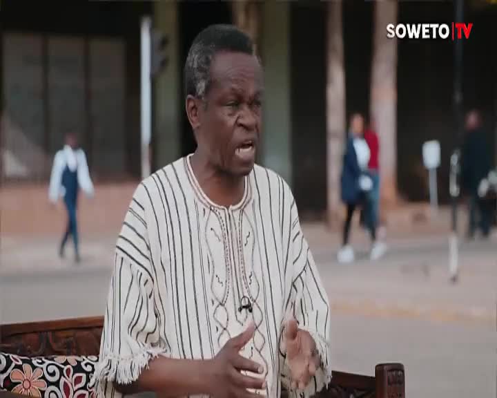 A man in a striped shirt gestures emphatically, speaking passionately on Soweto TV. Behind him, people casually stroll on a paved area, oblivious to the broadcast.
