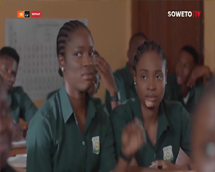 Two young women in matching green shirts sit at desks, their faces turned toward something off-screen, likely a lesson on Soweto TV. The one on the right is gesturing with her hand, while the other looks on with a slight frown.
