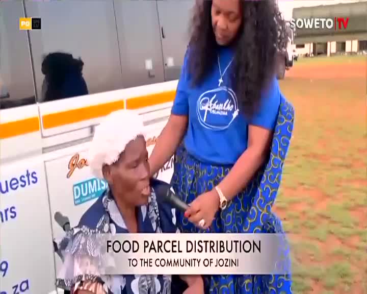 A woman in a blue shirt holds a microphone towards an elderly woman in a wheelchair, who is speaking. Soweto TV's logo is visible as the words "Food Parcel Distribution" are displayed on the screen.
