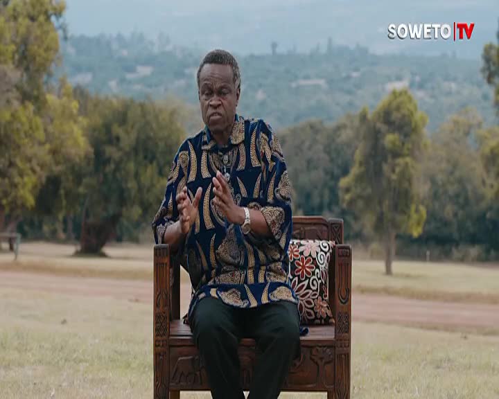 A man in a patterned shirt gestures with his hands, speaking on Soweto TV. He sits on a carved wooden bench, the green landscape of South Africa providing a backdrop.
