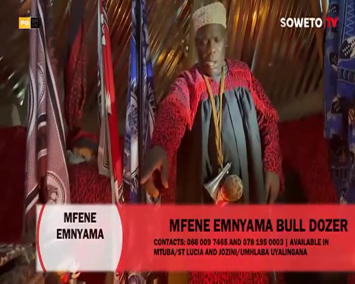 A man in traditional South African attire, adorned with a red and black patterned robe, stands before a backdrop of colorful flags. He gestures with his right hand, a small horn and a spherical object hanging from a cord around his neck.