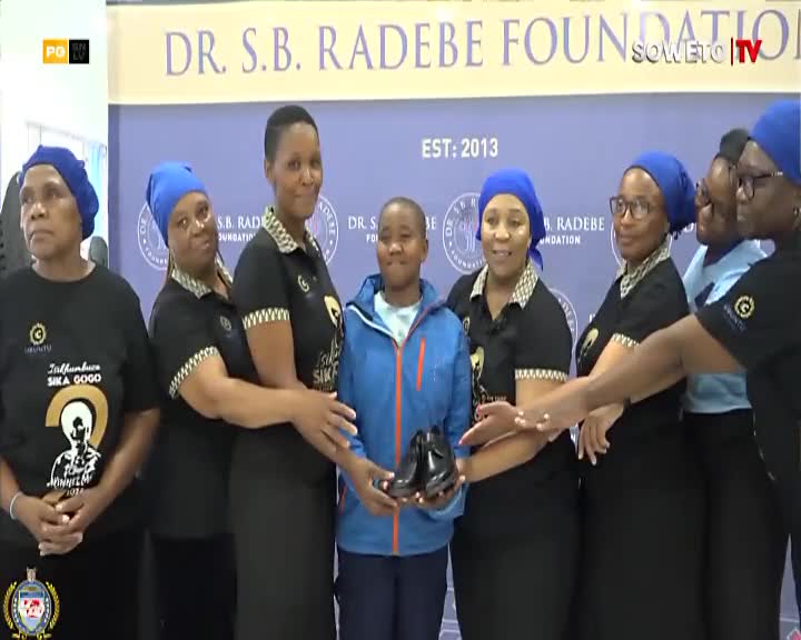 A young person stands center, holding a pair of shiny black shoes. A group of women, some in matching black tops with gold trim, surround them, their faces turned towards the child. The Dr. S.B. Radebe Foundation banner hangs behind them, with the Soweto TV logo visible.