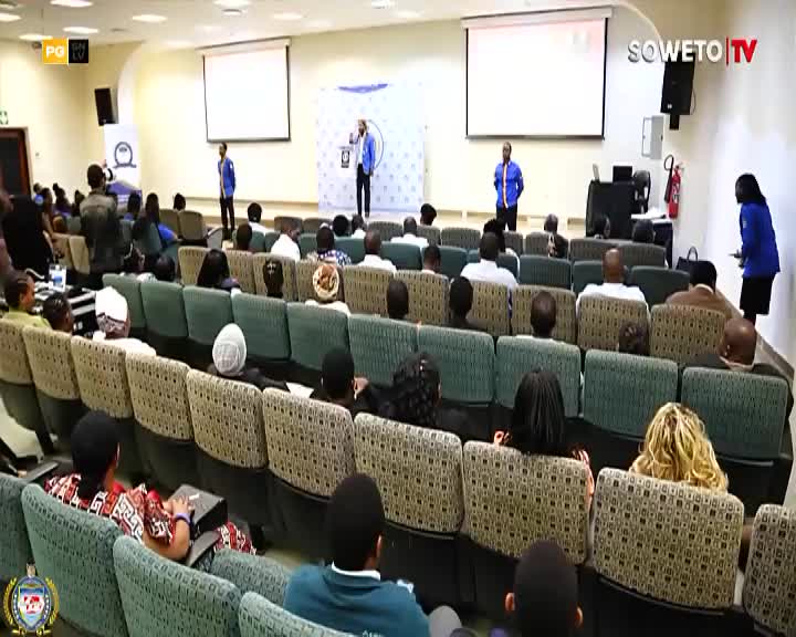 A man in a blue shirt addresses a seated audience in a conference room, with Soweto TV branding visible. Another person in a blue shirt stands near the podium, while a woman in a blue jacket walks away from it. A man in a blue shirt addresses a seated audience in a conference room, with Soweto TV branding visible. Another person in a blue shirt stands near the podium, while a woman in a blue jacket walks away from it.
