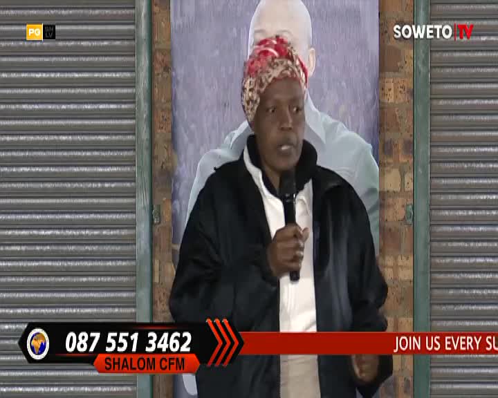 A woman in a patterned headscarf holds a microphone, speaking into it with a serious expression. Behind her, a large poster shows a man's silhouette, and a Soweto TV logo is visible.