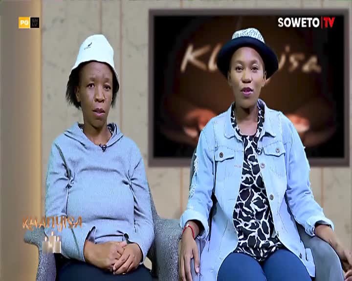 Two women sit on chairs, facing forward. The woman on the left wears a grey hoodie and a white bucket hat, while the woman on the right has on a denim jacket over a patterned top and a fedora.