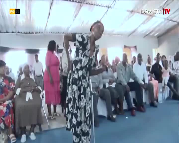 A woman in a black and white patterned dress leans on a crutch, her body angled towards a seated audience. The Soweto TV logo is visible in the upper right corner. A woman in a black and white patterned dress leans on a crutch, her body angled towards a seated audience. The Soweto TV logo is visible in the upper right corner.