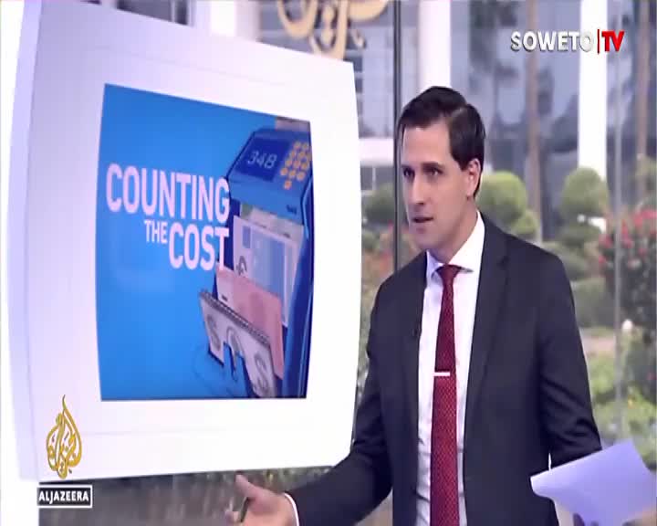 A man in a suit gestures while speaking, a screen behind him displays "COUNTING THE COST" with images of currency. Soweto TV's logo is visible in the corner. A man in a suit gestures while speaking, a screen behind him displays "COUNTING THE COST" with images of currency. Soweto TV's logo is visible in the corner.