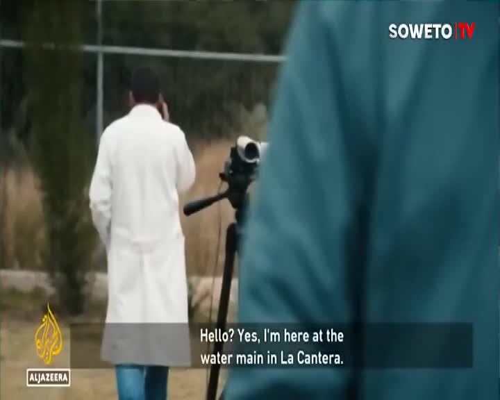 A man in a white lab coat walks away from the camera, speaking on his phone. A camera on a tripod stands nearby, ready for filming. A man in a white lab coat walks away from the camera, speaking on his phone. A camera on a tripod stands nearby, ready for filming.