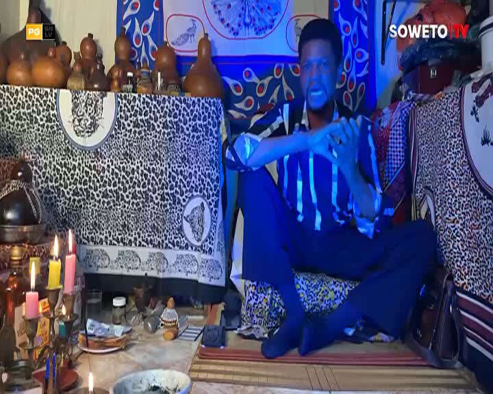 A man in a striped shirt sits cross-legged, his hands clasped in front of him, while colorful fabrics and gourds surround him. The Soweto TV logo is visible in the corner of the screen. A man in a striped shirt sits cross-legged, his hands clasped in front of him, while colorful fabrics and gourds surround him. The Soweto TV logo is visible in the corner of the screen.
