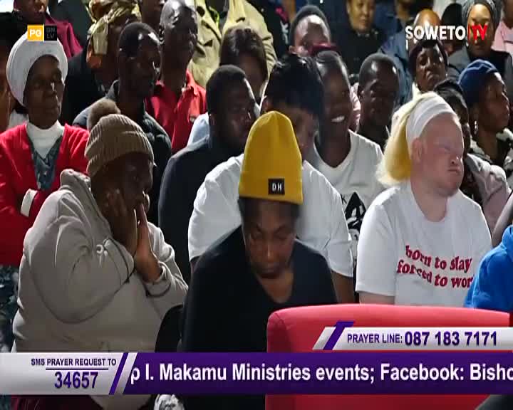 A crowd gathers, their faces turned upwards in anticipation. A woman in a yellow beanie bows her head, her hands clasped near her chin.