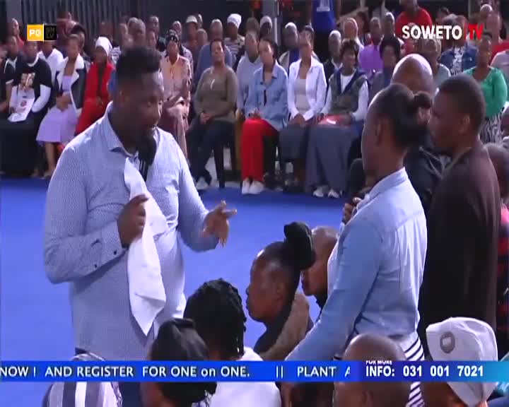 A man in a blue patterned shirt gestures with a white cloth in his hand, speaking to a crowd gathered in Soweto. The Soweto TV logo is visible in the background, with people seated and standing around the speaker.