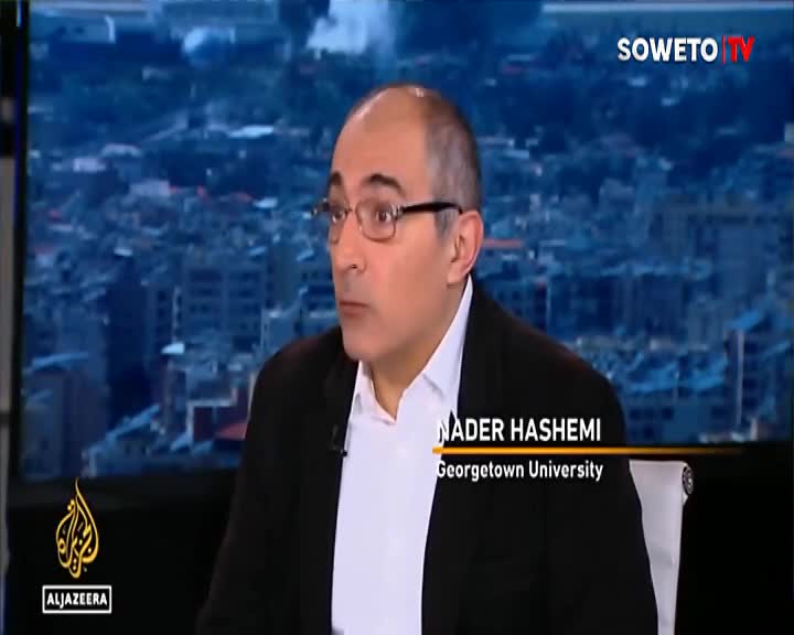 A man with glasses speaks on a Soweto TV broadcast, his name and affiliation displayed below him. Behind him, a cityscape appears on a screen, hinting at the topic of discussion.