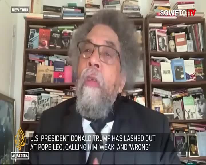 A man with a full grey beard and afro speaks animatedly, his glasses perched on his nose. Bookshelves packed with titles line the wall behind him, some with prominent faces on their covers.